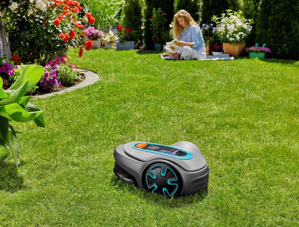Image de la tondeuse Gardena Sileno avec une femme qui fait du jardinage en arrière plan.